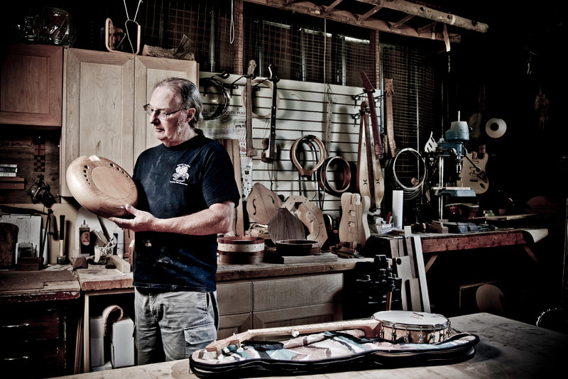 A man in a dark t-shirt stands in a workshop. He's holding the circular body of a custom made banjo.