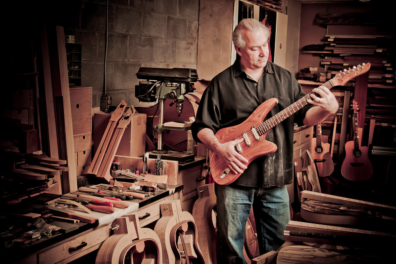 A man stands in a woodworking shop, pretending to play one of the guitars he has made.