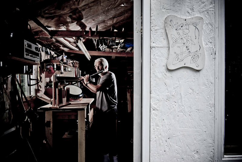 A white-haired man stands within a small workshop. Illuminated with bright lights, he plays an Irish flute that he made.
