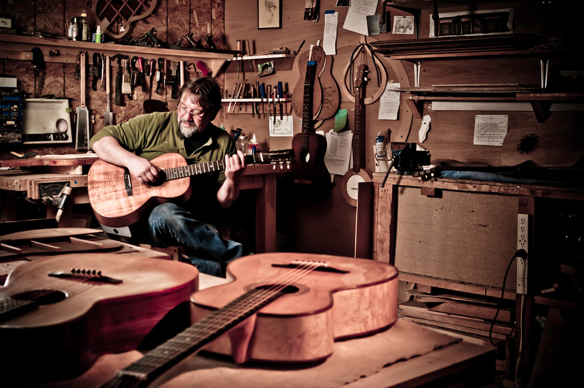 A bearded man plays his favourite, hand-made guitar in his workshop. More guitars rest on a table near the camera.