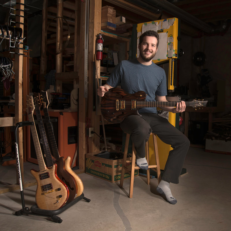 A young bearded man sits on a stool. Smiling at the camera, he holds a beautiful hand-made guitar.