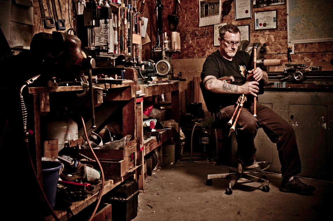 A man sits on a stool in the corner of a cozy workshop where he plays a vintage Irish bagpipe set.