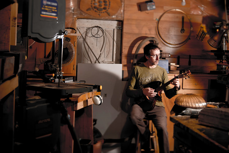 A bespectacled man sits on a stool in a tiny workshop. He plays a custom built mandolin as a small light illuminates him.