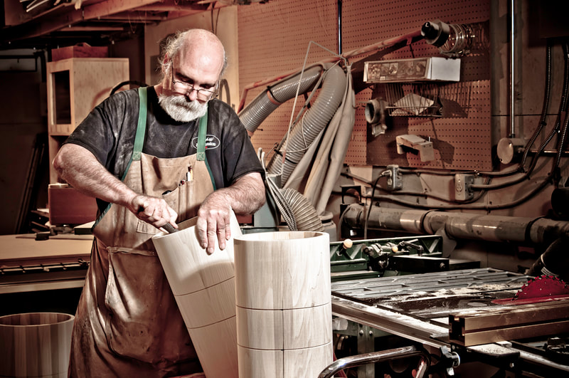 A bearded, balding man stands in his workshop. With tool in his hand, he fine tunes a new drum shell.