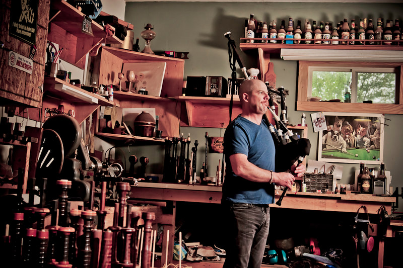 A man in a blue shirt stands in his small workshop while playing the highland bagpipes that he made.